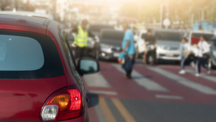 Fototapeta premium Rear side view of red car brake on the road. Traffic police are directing cars to stop at a crosswalk where young children are passing by. Blurry showing a line of cars parked on the opposite side.