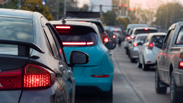 Rear side view of luxury car with turn on brake light. Many cars parked in a queue on concrete road. There's a long line of traffic. Blurred of traffic red light conctrol and city with trees.