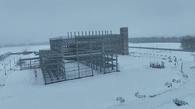 Massive concrete framework rises across snowy industrial territory. Temporary structures and hangars reinforce raw construction atmosphere
