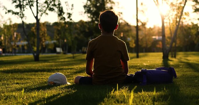 Kid boy relaxes on lawn at sunset. Silhouette of a child in an outdoor park during golden hour. Summer park scene suggests break after children's play. There is cap and backpack next to child.