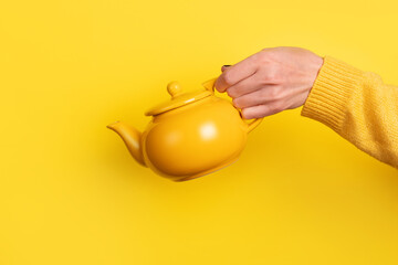 A hand in a yellow sweater holds a yellow teapot against a vibrant yellow background, creating a bold and modern monochromatic composition.