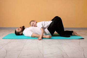 Young girl resting on her father&rsquo;s chest as they lie on a blue yoga mat in a minimal, home setting with warm tones.
