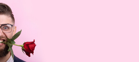 Playful man with glasses holds a red rose in his mouth against a pastel pink background, perfect for love, romance, or humorous concepts. panoramic layout