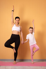 Smiling mother and daughter in activewear performing yoga tree pose together on a pink mat against a peach background.