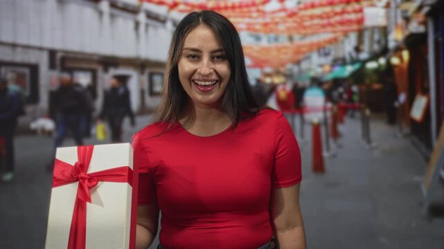 Woman holding gift box on a busy street market smiling with open mouth and outstretched arm; holiday giving joy.