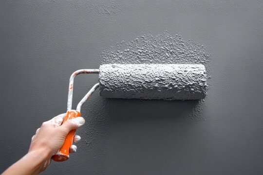 hand holding orange paint roller applying thick grey paint to a wall, textured wet stroke conveying focused DIY satisfaction