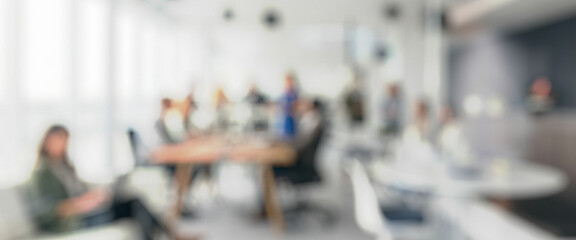 Blurred view of a business meeting in a modern office. People are seated around a large table, discussing ideas while several monitors display information.