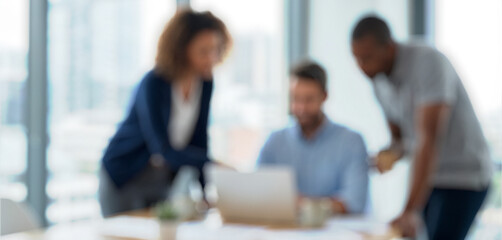 Three professionals collaborate in a modern office. One person gestures while another looks at a laptop, all seated at a conference table with a city view behind them.