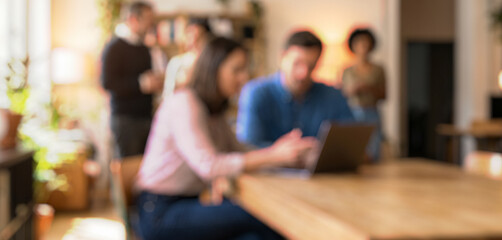 Blurred image of a person using a laptop in a modern workspace. Individuals collaborate in the background, showcasing teamwork in a comfortable environment.