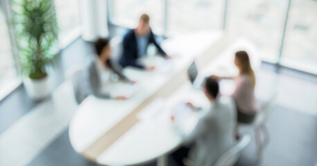 Blurred image of a business meeting in a modern office. Professionals gather around a circular table discussing projects, with sunlight streaming through large windows.
