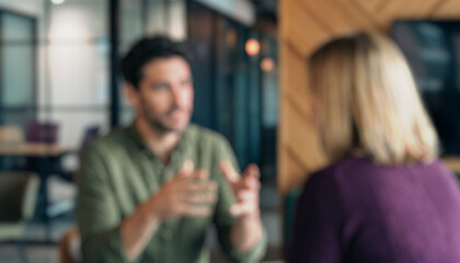 Blurred image depicting two individuals engaged in a conversation indoors. One person gestures while the other listens attentively in a modern office environment.