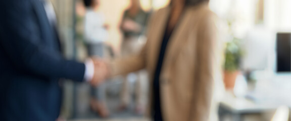 Blurred image of two people shaking hands in an office. In the background, additional individuals are engaged in discussion. The setting appears professional and collaborative.