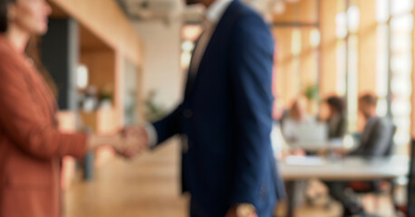 Blurred image of two people shaking hands in an office environment with people working at desks in the background. Natural light illuminates the modern workspace.