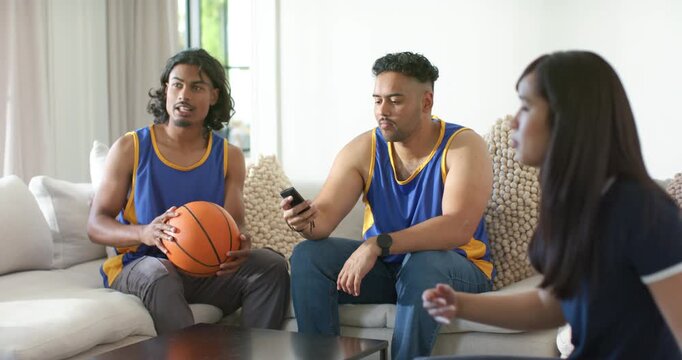 African American friends on sofa in jerseys holding basketball  and  remote pointing at TV woman mov