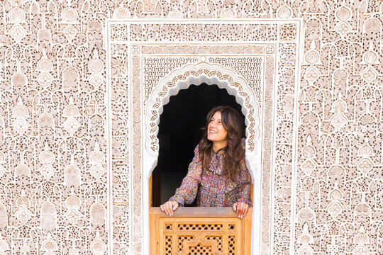 A woman in a floral shirt looks out from an ornate window with intricate carvings. Madrasa di Ben Youssef,Marrakech,Morocco
