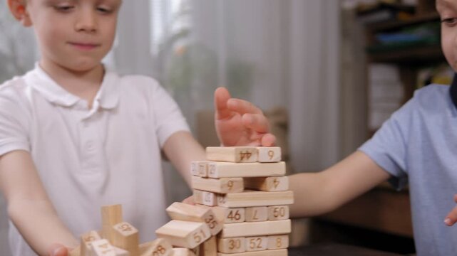 Close-up of two emotional school-age boys playing an exciting board game with wooden blocks. The boy takes out a wooden block from the tottering tower.