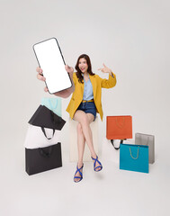 Mobile shopping app promotion with Asian woman holding blank smartphone screen among colorful shopping bags presenting ecommerce platform digital payment and modern retail marketing concept.