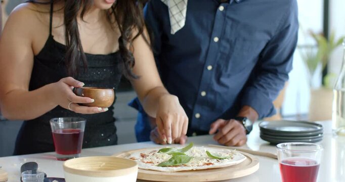 Asian woman and African American man adding basil leaves to pizza at counter, preparing to serve