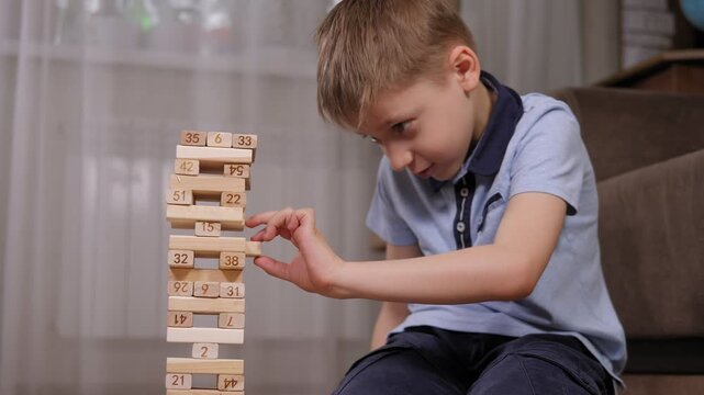 An excited emotional boy is playing a board game of jenga, he takes out one wooden block from a wobbly tower. Jenga logistic game for kids development.