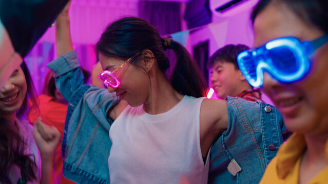 Closeup group multiethnic young adult Asia friend wear futuristic neon glowing glasses dance and drink alcohol beer fun hangout party in living room night indoor home. Disco lights illuminating house.
