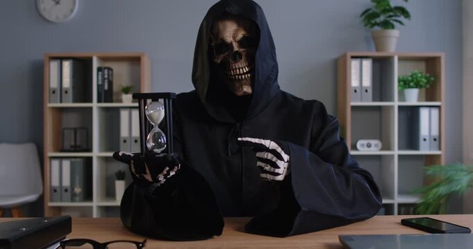 Sinister figure wearing dark hooded cloak and skull mask holding hourglass while pointing with skeletal finger at time. Grim reaper of death reminding of passing time and inevitable end of life.