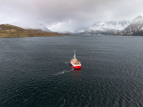Aerial view of a red and white boat cuts through the dark, rippling waters, framed by the snow-dusted mountains under a moody sky, Evenes, Nordland, Norway.