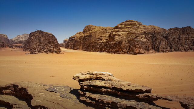 Aerial view of sun-scorched desert sands meet rugged, ancient rock formations under a vast, clear sky, Wadi Rum Village, Aqaba Governorate, Jordan.