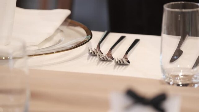 Close up of a hand carefully arranging silver forks on a white tablecloth next to a glass plate. Static shot