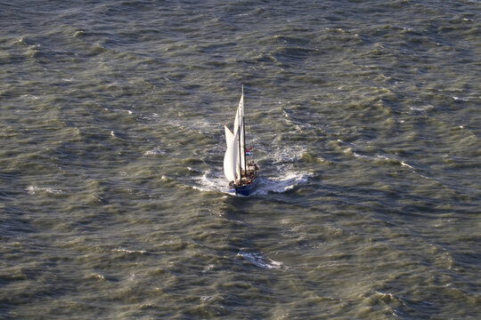 Hook of Holland, Netherlands - 11 February 2026: Aerial view of a sailboat cutting through the choppy, sun-kissed waters of the North Sea, its white sails contrasting against the dark waves.
