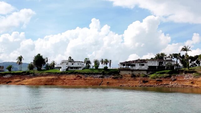Cinematic POV shot from a boat approaching the decaying Hacienda La Manuela, Pablo Escobar's notorious abandoned luxury estate on the shores of Guatape Reservoir, Colombia.
