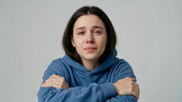 Portrait of a young sad woman crying with tears running down her cheeks. Upset depressed girl in blue hoodie looking at camera with despair. Mental health and grief concept