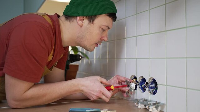 Concentrated male electrician in uniform using a screwdriver to install and repair a new electrical wall socket, working with wiring during a kitchen remodeling or construction project