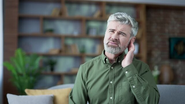Close-up of man touching his ear and wincing from sharp pain at home. Senior male with gray hair feeling discomfort, irritation, suffering from ear infection or inflammation, reacting to sudden ache
