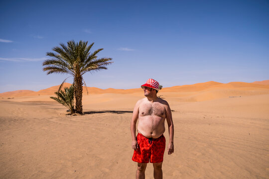 Overweight man with swimming shorts and hat standing in the desert of Morocco