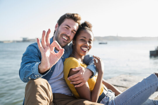Portrait of happy young couple sitting at the waterfront gesturing, Lisbon, Portugal