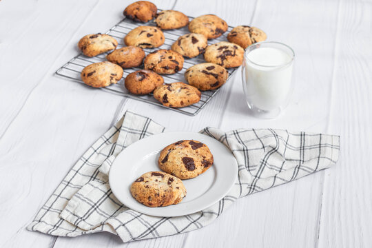 Fresh homemade chocolate chip cookies on a plate with milk and cooling rack on light wooden background