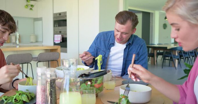 Family teen eating at kitchen table, man lifting pesto pasta from bowl, tasting for flavor coughing