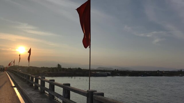 Patriotic Vietnam Bridge features long concrete roadway decorated with fluttering red flags during sunset. Scenic river crossing highlights national pride on Vietnam Bridge under golden evening sky.