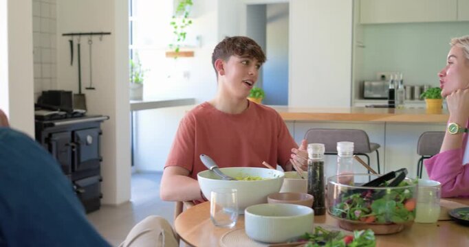 Teenage son sharing story at kitchen table over dinner, parent listening, reaching for salad bowl