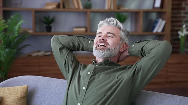 Close-up of smiling mature man relaxing with eyes closed and hands behind head at home. Happy male resting on sofa in cozy living room, enjoying peace, comfort, and pleasant relaxation after work