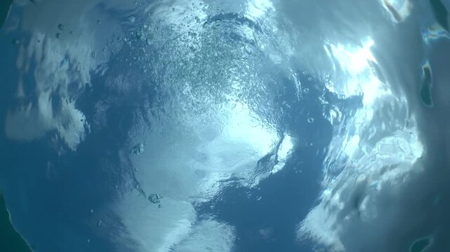 Camera points towards the surface of the ocean capturing the surface and a scuba diver's bubbles.