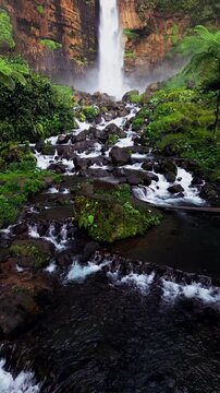 Vertical video at Kapas Biru Waterfall capturing white rushing water descending beside a massive limestone cliff with rich vegetation and untouched tropical jungle scenery.