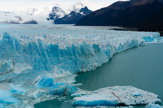 View of a massive, textured Perito Moreno Glacier meets the teal waters, framed by dark mountains and snowy peaks, creating a dramatic scene, El Calafate, Santa Cruz Province, Argentina.