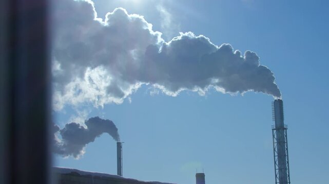 Smoke coming out of two tall factory chimneys against a blue sky. Air pollution.
