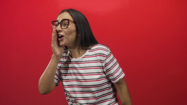 Woman cupping hand to mouth in red studio wearing striped shirt and glasses; cheerful secret sharing surprise.