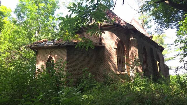 Old, abandoned brick building in the gothic style, likely a chapel or a church, slowly being reclaimed by nature as lush green vegetation grows wild and unchecked all around its weathered walls