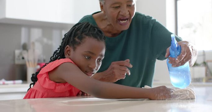 Sr African-American grandma spraying bottle, guiding granddaughter wiping kitchen counter with rag