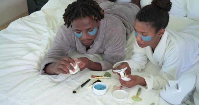 Couple spotting products on bed, in robes with eye patches, testing LED, inspecting mask for facial