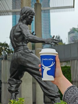 Feb 12, 2026 - Hong Kong, China: A photorealistic image juxtaposes Bruce Lee's bronze statue with a Luckin Coffee cup in a Hong Kong plaza.