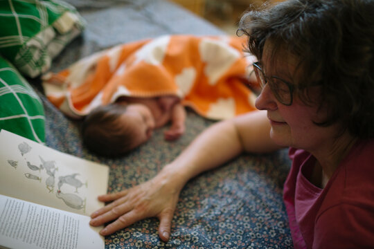 Senior woman reading storybook next to grandson sleeping on sofa at home